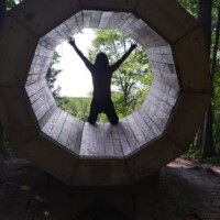 Silhouetted figure inside octagonal wooden sculpture at Agnes Andreae Nature Preserve Michigan — arms raised, forest light framing the structure