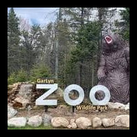 Entrance sign for Garlyn Zoo Wildlife Park in Naubinway, Michigan, with a large bear statue beside the sign and tall trees in the background.