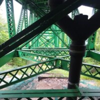 Underside view of a green steel bridge with triangular trusses and cross‑bracing, featuring a large black pipe with a Y‑junction in the foreground, above rocky ground and surrounded by trees.
