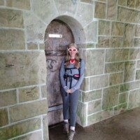 Person standing in front of a small arched wooden door set into a stone wall at Cut River Bridge, wearing a gray outfit, backpack, and red‑tinted sunglasses.