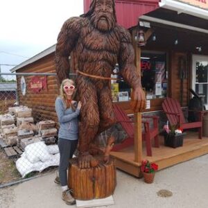 Posing with a carved wooden Bigfoot statue outside Gustafson’s Smoked Fish in Moran, Michigan, wearing heart‑shaped sunglasses for a playful roadside photo.