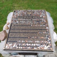 Memorial plaque dedicated to Heath Michael Robinson, mounted on a stone base with coins and small rocks placed on top as tokens of respect, surrounded by grass.