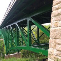 Side view of the green steel truss structure of Cut River Bridge, showing interconnected triangular beams above a wooded gorge with a stone support pillar in the foreground.