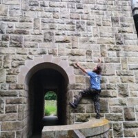 Person pretending to climb a stone wall with an arched passageway at Cut River Bridge, wearing a blue shirt and dark pants, using both hands and feet to ascend.