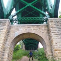 Person standing on a dirt path beneath a large green steel bridge supported by stone arches, with intricate metalwork overhead and greenery surrounding the base.