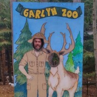 Painted photo board with a safari explorer and large deer illustration, featuring cutouts for faces, with a person posing in the explorer cutout and stylized trees in the background and a GarLyn Zoo Banner above head.