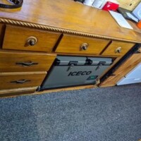 Converted bus kitchen with white stove in background, wooden dresser in foreground containing ICECO portable refrigerator behind open compartment, on carpeted floor.