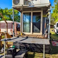 Open back deck attached to converted bus with metal railing, small table, storage bins, and sliding glass doors leading into the interior.