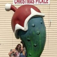 A large green pickle sculpture wearing a Santa hat labeled “Christmas Pickle,” with two people posing in front—captured at the World’s Largest Christmas Pickle display in Fort Smith, Arkansas.