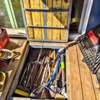 Open deck storage compartment on converted bus containing hand tools including broom, hammer, hacksaw, crowbar, pipe wrench, and other tools organized in wooden enclosure