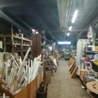 Interior of Refab St Louis Repurpose Shop showing a densely packed workshop area with reclaimed wood, PVC pipes, shelving units, and miscellaneous salvage materials arranged in a cluttered but resource-rich environment.
