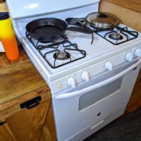 Full-size white gas stove in converted bus kitchen with frying pan, covered pot, wooden cabinets, and countertop with yellow bottle, all set against a carpeted floor.