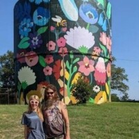 Two people stand in front of a tall cylindrical water tower painted with a vibrant floral mural featuring daisies, leaves, and a bee, set against a clear blue sky.