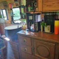 Bus kitchen interior with wooden drawers, countertop appliances including coffee maker, open shelves with jars, and white curtain above small sink under window