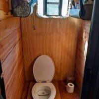 Compact bathroom inside converted bus with wood-paneled walls, open composting toilet, small window, and shelves holding toiletries and a metal container.