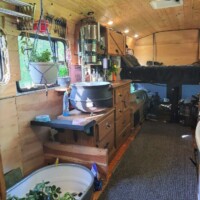 Converted bus kitchen with wooden cabinetry, sink under curtained window, produce basket, countertop appliances, and a view into built-in shelving and sleeping loft above driver’s area.