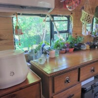 Converted bus interior with wooden dresser topped with potted plants and decor, ICECO refrigerator below, air conditioning unit above curtained window, and hanging net organizer on right wall.
