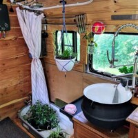 Bus kitchen sink area with black basin, silver faucet, wooden cabinetry, hanging plant, pink soap dish, and window with white curtain above small indoor tub containing potted plants.