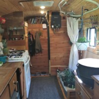 Interior of converted bus kitchen with full-size white stove, wood-paneled walls and ceiling, countertop with produce, bookshelves above, and potted plants beside sink and window