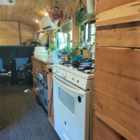 Converted bus kitchen with full-size white stove, wood countertops, overhead cabinets with baskets, hanging plants by curtained windows, and wood-paneled ceiling.