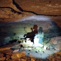 Inside War Eagle Cavern Arkansas, light casts the silhouette of a seated figure drinking onto rough cave walls, blurring shadow and stone.