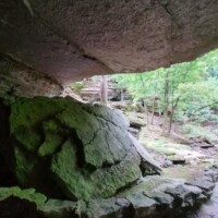 Cracked green‑tinged boulder rests beneath an overhanging rock slab, forming a partial cave in a lush forest of layered cliffs and trees at War Eagle Caverns Arkansas