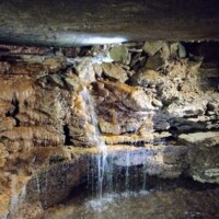 Inside War Eagle Cavern Arkansas, a dim waterfall spills over slick rock into a shallow pool, caught in a single beam of light.