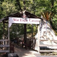 Rustic wooden gateway marked “War Eagle Cavern Arkansas,” with a tipi tucked just beyond, all framed by dense trees like it’s guarding the cave’s secrets.