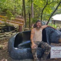 At War Eagle Cavern Arkansas, a bearded visitor lounges on a black bear‑shaped bench beside a rock waterfall, camo pants blending into the wooded backdrop.