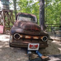 Rust‑pitted vintage truck with mannequin legs poking out from underneath, “Redneck Work Ethic” plate, and a Hillbilly Hollar water tower in the trees behind.