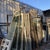 Outdoor display at Refab St Louis Repurpose Shop showing a variety of reclaimed window frames and panes stacked against a metal building beneath a hanging “WINDOWS” sign, highlighting salvage materials available for creative reuse and DIY renovation.