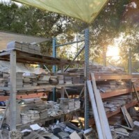 Outdoor shelving area at Refab St Louis Repurpose Shop featuring multiple stacks of tiles or stone slabs arranged on metal and wood racks beneath a canopy, with trees and filtered sunlight in the background, showcasing reclaimed materials for reuse and renovation.