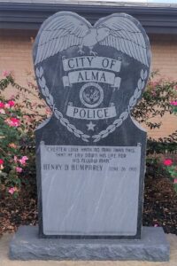 Henry D. Humphrey memorial, Stone, shaped with a heart-like top and engraved eagle. Center badge emblem encircled by laurel wreath. Inscription reads: “Greater love hath no man than this, that he lay down his life for his fellow man.” Surrounded by blooming rose bushes, placed in front of a brick wall. Honors a fallen Alma, Arkansas police officer killed in the line of duty.