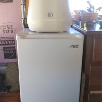 White Lomi countertop composter placed on top of a small white freezer in converted bus kitchen, next to wooden cabinet and window with potted plant.