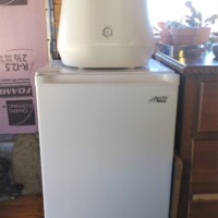 White Lomi countertop composter placed on top of a small white freezer in converted bus kitchen, next to wooden cabinet and window with potted plant.