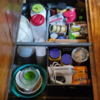 Open floor cabinet in converted bus containing stacked bowls, jars, small kitchen appliances, packaged foods, plastic containers, and rolled paper products