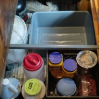 Organized under-compartment bus storage with various plastic containers, mason jar, white jug, packaged food, and cleaning supplies inside black bin and gray tub