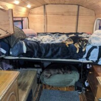 Converted bus living area with elevated bed above wooden cabinets, one dog on the bed, one dog in a bed below, wood-paneled ceiling with recessed lighting, and carpeted floor