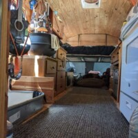Converted bus kitchen with wood-paneled ceiling, carpeted floor, hanging black pot, wooden cabinetry on both sides, and compact white appliance beside drawers