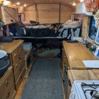 Converted bus kitchen featuring full-size propane range and cast iron cauldron sink set into wooden countertop beneath curtained window.