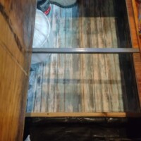 Under-floor storage compartment in converted bus revealed by lifted wood panel, containing shoes, helmet, and black cylindrical object, with striped surface inside