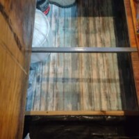 Under-floor storage compartment in converted bus revealed by lifted wood panel, containing shoes, helmet, and black cylindrical object, with striped surface inside