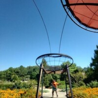 Large metal sculpture with circular frames and wire extensions resembling abstract antennae at the Botanical Garden of the Ozarks, with a person standing beneath it in a garden bed of orange and yellow flowers under a clear blue sky