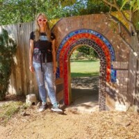 Small arched mosaic doorway embedded in a wooden fence at the Botanical Garden of the Ozarks, decorated with colorful tiles in red, blue, and black, with a person in sunglasses, black shirt, jeans, and sneakers standing beside it on a dirt path, surrounded by greenery and trees