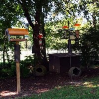 Four colorful birdhouses mounted on tall wooden poles in a shaded garden area at the Botanical Garden of the Ozarks, with hollowed logs below and a fenced shed in the background