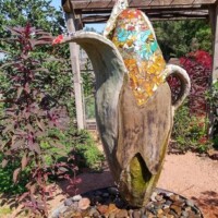 Large outdoor sculpture of a corn cob with partially peeled husks at the Botanical Garden of the Ozarks, featuring colorful mosaic tiles in yellow, orange, blue, and white, placed on a bed of rocks with surrounding greenery and a wooden pergola in the background