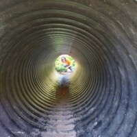 Interior view of a large corrugated metal tunnel at the Botanical Garden of the Ozarks, with concentric ridges creating a dramatic pattern leading to a person in a red shirt crouching at the far end, surrounded by greenery and natural light