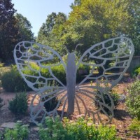 Large metal butterfly sculpture with lattice-patterned wings placed in a garden bed at the Botanical Garden of the Ozarks, surrounded by green shrubs and plants with trees and a clear blue sky in the background