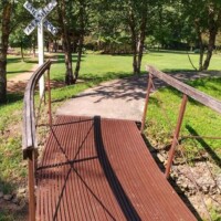 Small wooden bridge with metal railings positioned as part of the vintage train sculpture display at the Botanical Garden of the Ozarks, crossing a shallow ditch and leading to a railroad-style sign reading ‘RAIL SING’ and ‘XING,’ surrounded by green grass and trees
