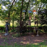 Seven bird feeders of various designs and colors hanging from poles in a wooded garden area at the Botanical Garden of the Ozarks, with mulch-covered ground and a decorative pedestal planter on the right